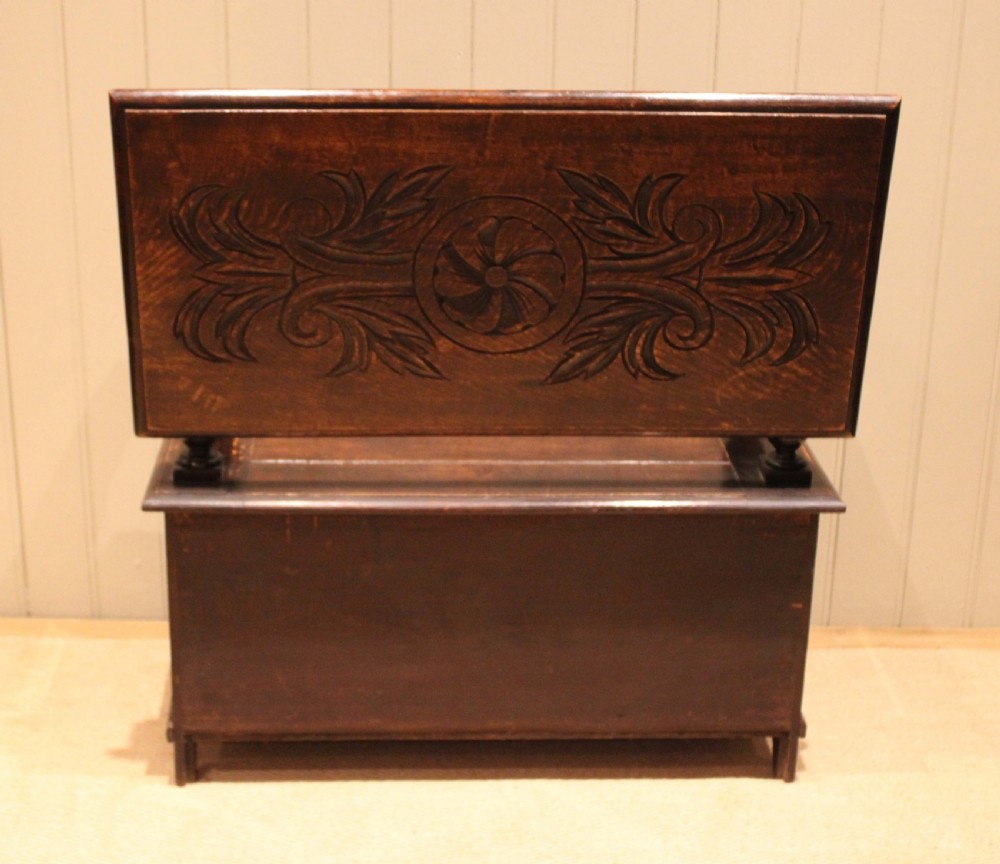 A carved dark oak monks bench featuring a relief-carved backrest, turned arm supports, and a deep box chest storage base. View XIII