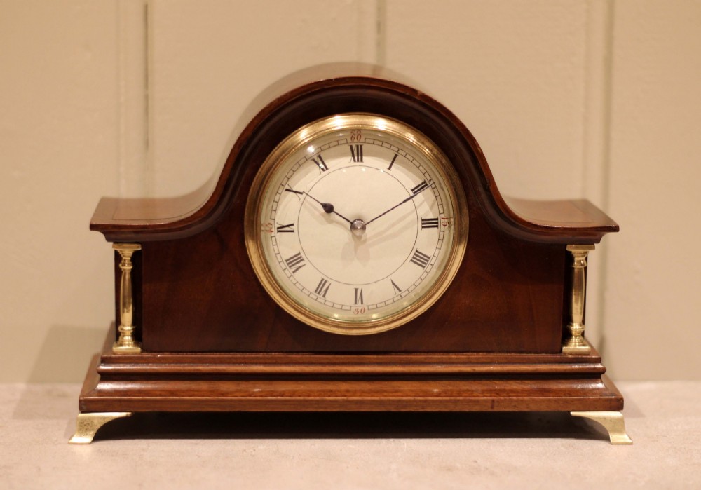 An Edwardian mahogany mantel clock featuring an arched top, turned brass side columns, brass feet, and a white dial with Roman numerals.