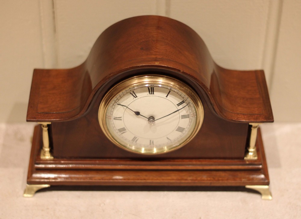 An Edwardian mahogany mantel clock featuring an arched top, turned brass side columns, brass feet, and a white dial with Roman numerals. View VI