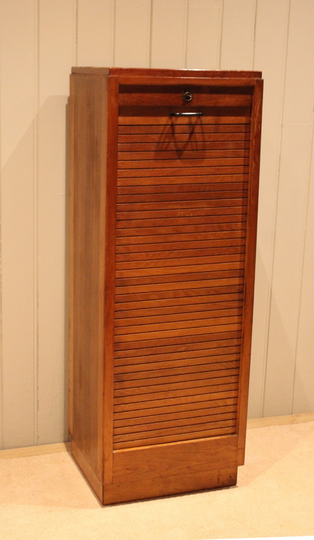 A tall, narrow antique oak filing cabinet with a vertical slatted tambour shutter front, top metal pull handle, and keyhole lock.