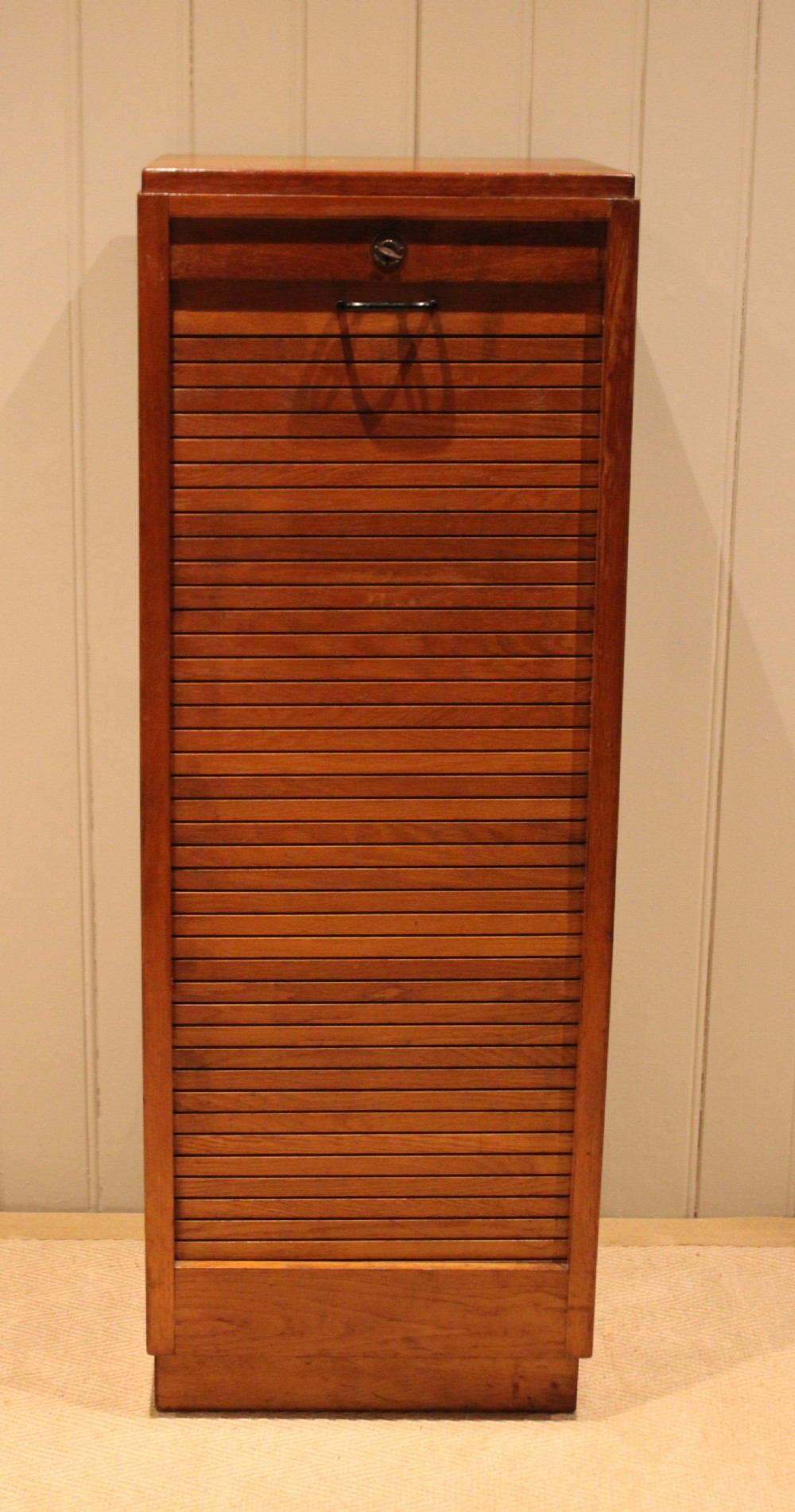A tall, narrow antique oak filing cabinet with a vertical slatted tambour shutter front, top metal pull handle, and keyhole lock. View III
