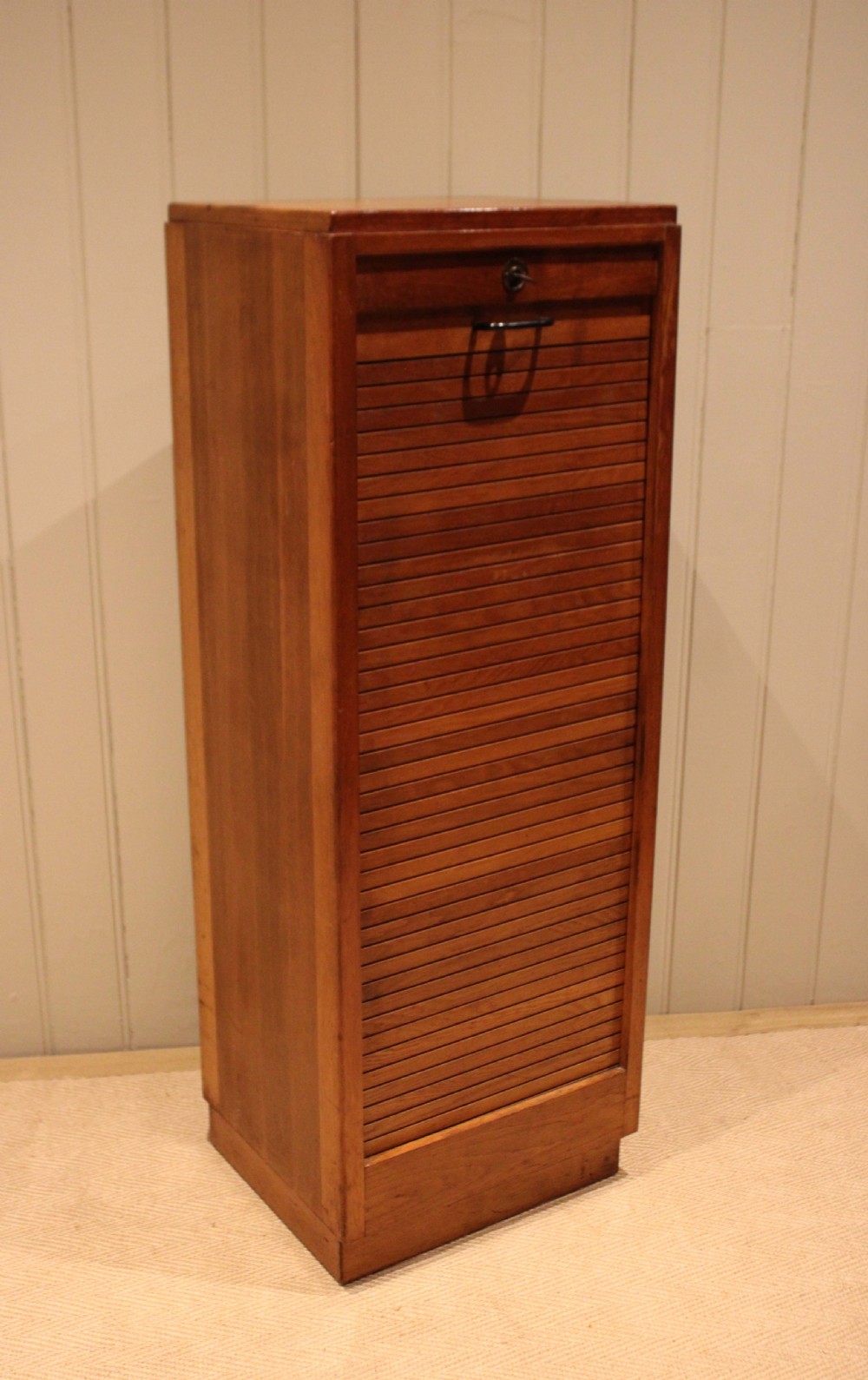 A tall, narrow antique oak filing cabinet with a vertical slatted tambour shutter front, top metal pull handle, and keyhole lock. View VII