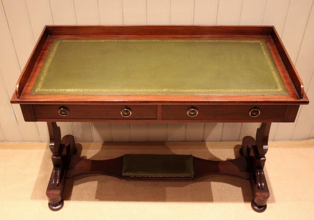 A mahogany writing table featuring a green gilt-tooled leather inset writing surface, a three-sided gallery rail, two drawers with brass ring pull handles, and a trestle base with a central footrest.
