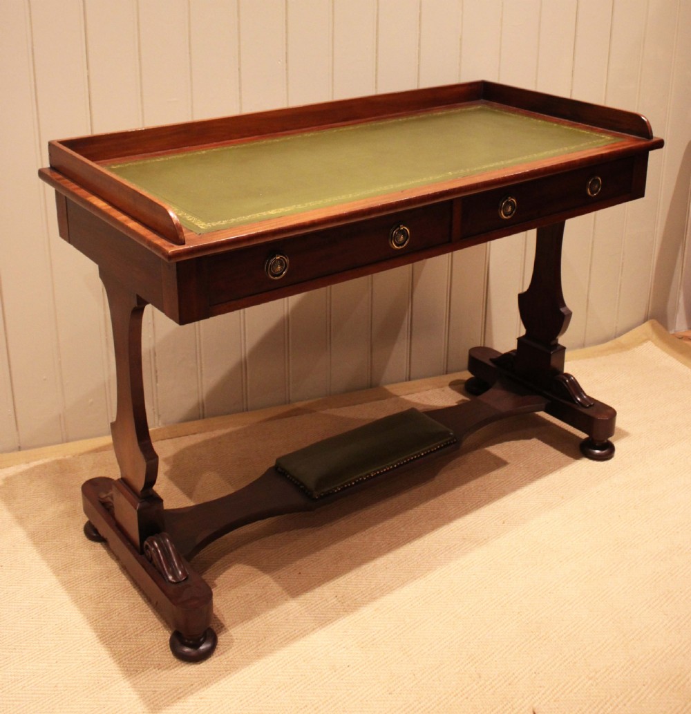 A mahogany writing table featuring a green gilt-tooled leather inset writing surface, a three-sided gallery rail, two drawers with brass ring pull handles, and a trestle base with a central footrest. View II