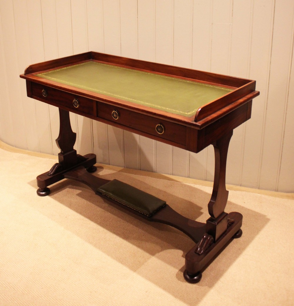 A mahogany writing table featuring a green gilt-tooled leather inset writing surface, a three-sided gallery rail, two drawers with brass ring pull handles, and a trestle base with a central footrest. View III