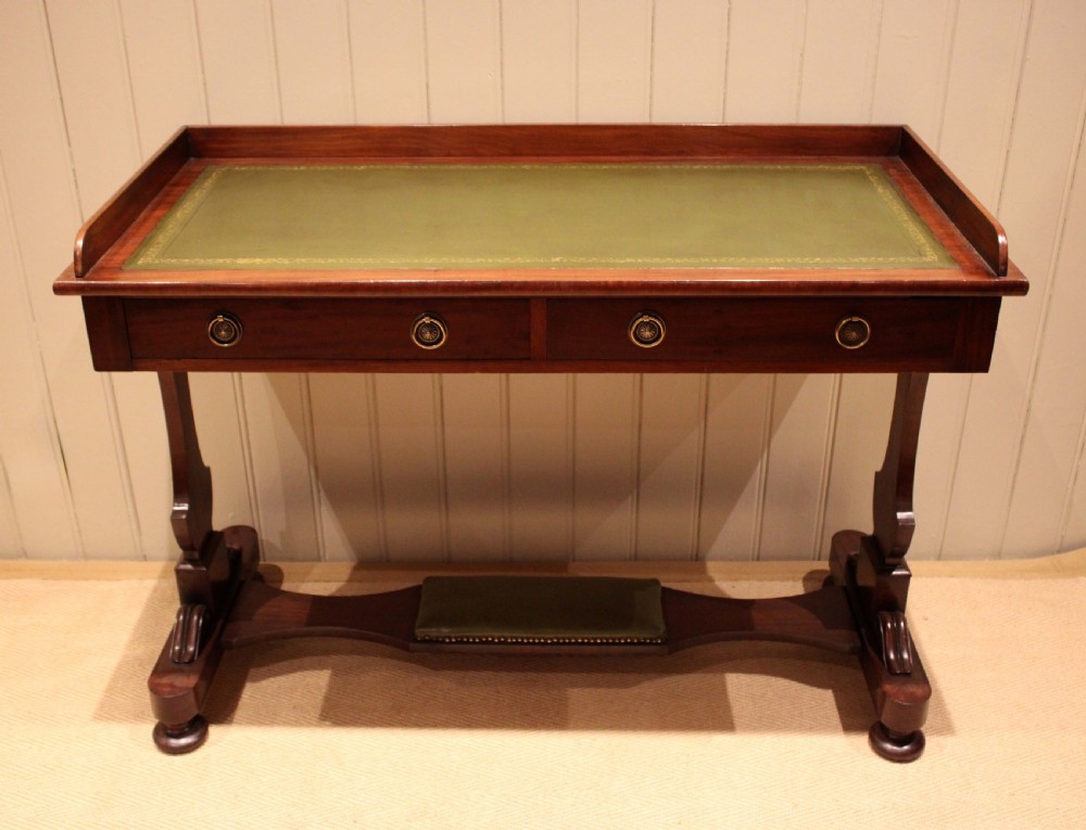 A mahogany writing table featuring a green gilt-tooled leather inset writing surface, a three-sided gallery rail, two drawers with brass ring pull handles, and a trestle base with a central footrest. View IV