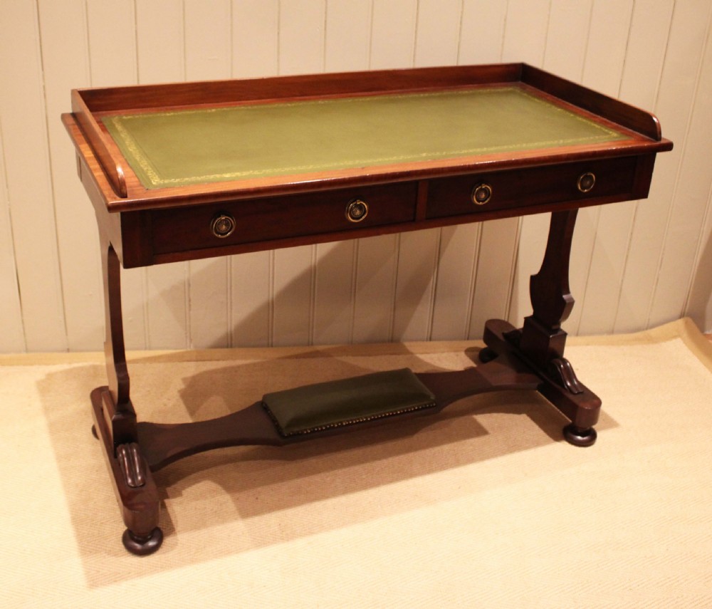 A mahogany writing table featuring a green gilt-tooled leather inset writing surface, a three-sided gallery rail, two drawers with brass ring pull handles, and a trestle base with a central footrest. View V