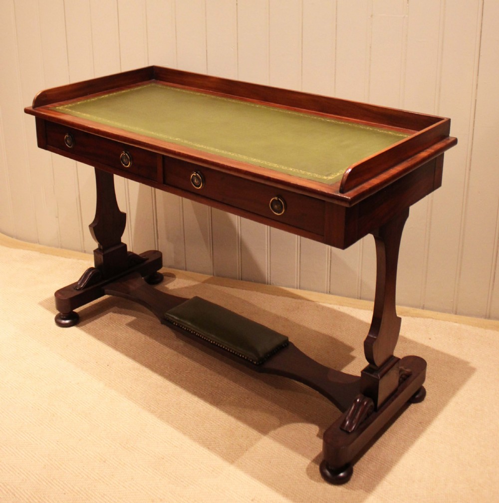 A mahogany writing table featuring a green gilt-tooled leather inset writing surface, a three-sided gallery rail, two drawers with brass ring pull handles, and a trestle base with a central footrest. View VI