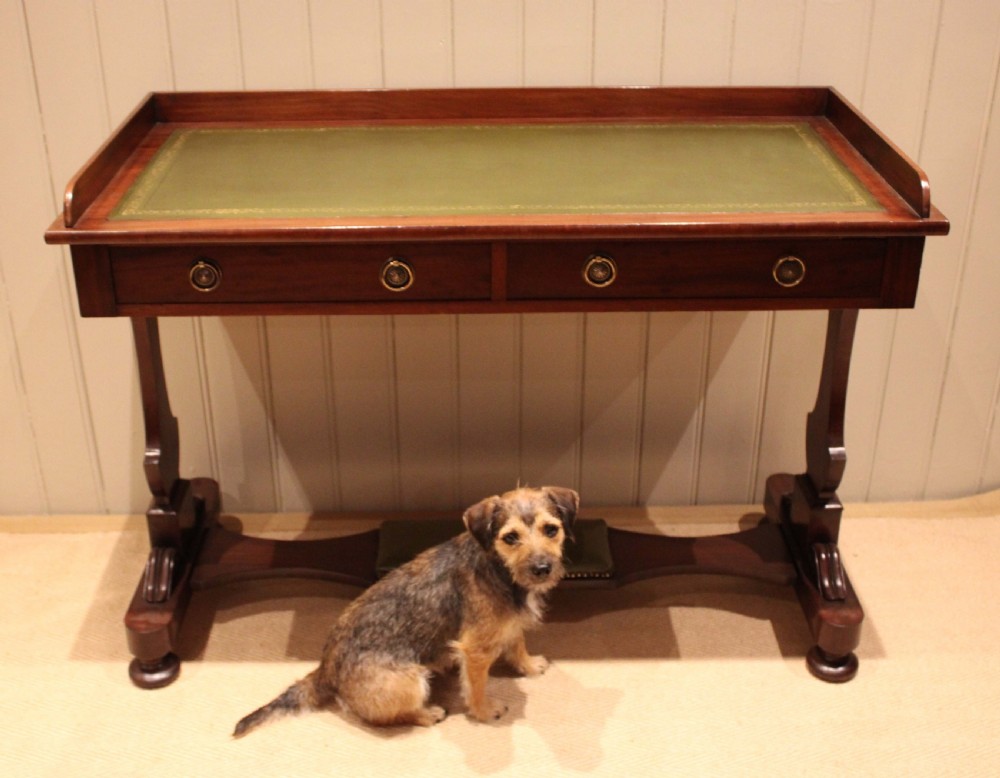 A mahogany writing table featuring a green gilt-tooled leather inset writing surface, a three-sided gallery rail, two drawers with brass ring pull handles, and a trestle base with a central footrest. View VII