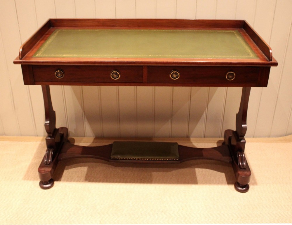 A mahogany writing table featuring a green gilt-tooled leather inset writing surface, a three-sided gallery rail, two drawers with brass ring pull handles, and a trestle base with a central footrest. View VIII