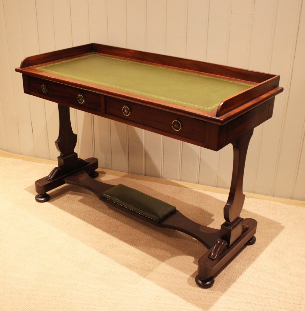 A mahogany writing table featuring a green gilt-tooled leather inset writing surface, a three-sided gallery rail, two drawers with brass ring pull handles, and a trestle base with a central footrest. View IX