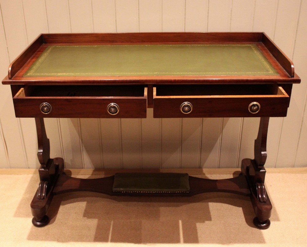 A mahogany writing table featuring a green gilt-tooled leather inset writing surface, a three-sided gallery rail, two drawers with brass ring pull handles, and a trestle base with a central footrest. View X