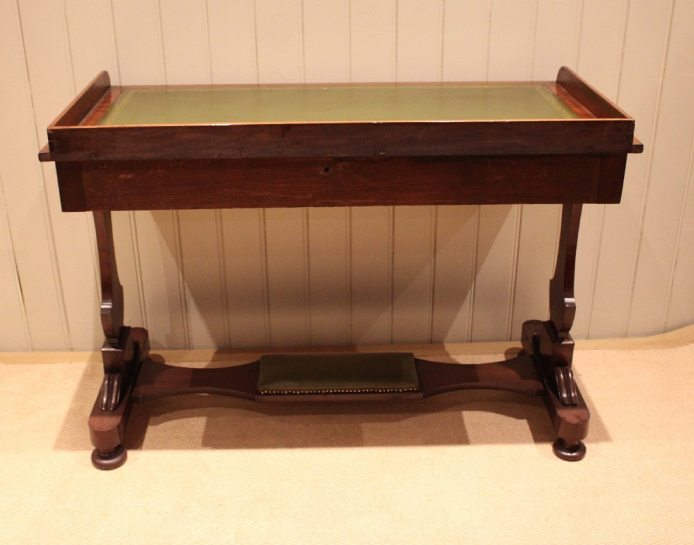 A mahogany writing table featuring a green gilt-tooled leather inset writing surface, a three-sided gallery rail, two drawers with brass ring pull handles, and a trestle base with a central footrest. View XII