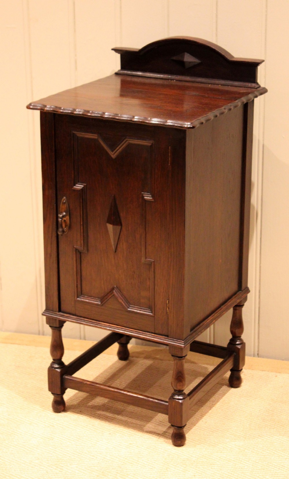A dark oak bedside cabinet with a geometric panelled door, turned legs, low stretchers, and an arched gallery back.