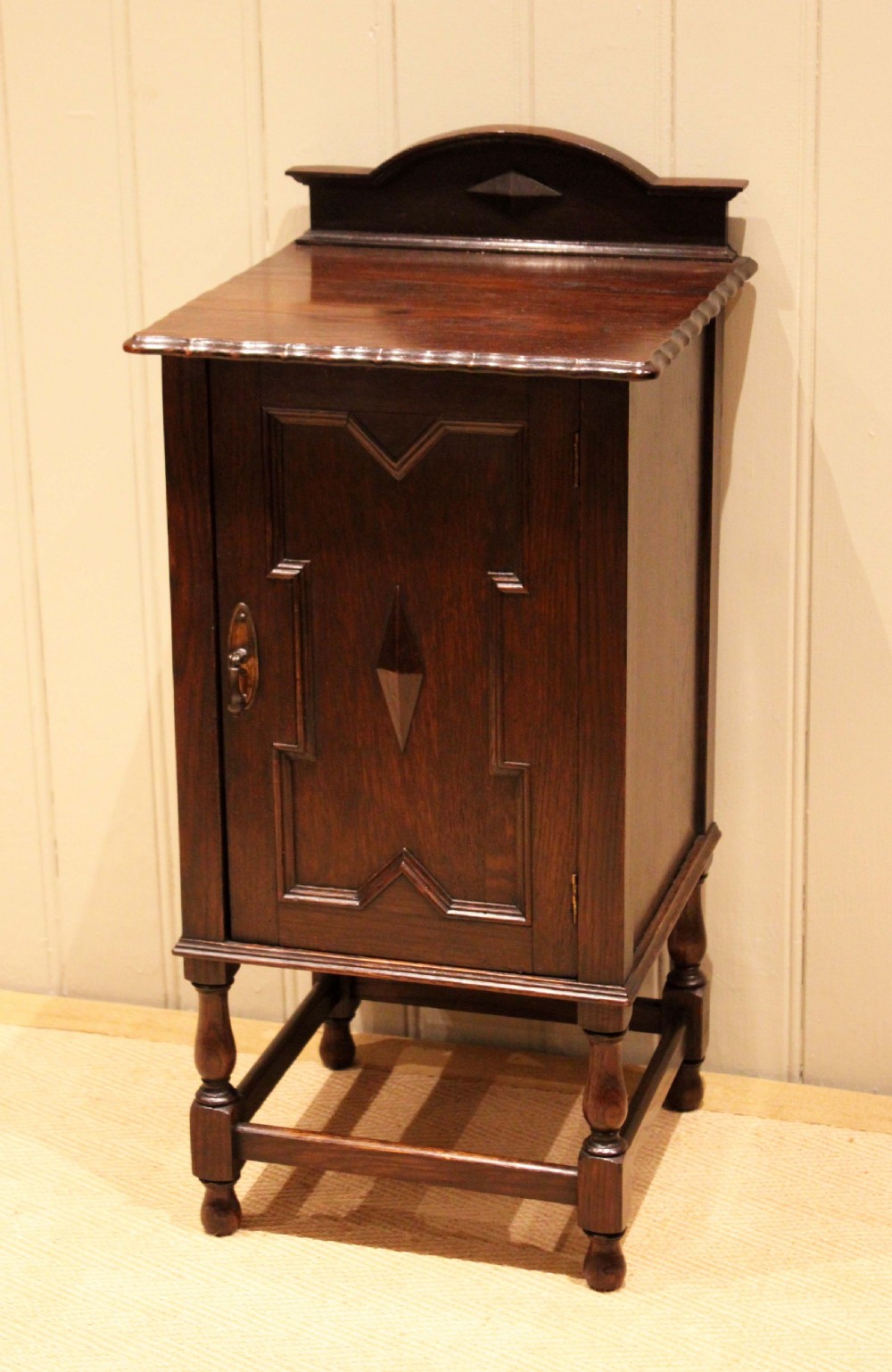 A dark oak bedside cabinet with a geometric panelled door, turned legs, low stretchers, and an arched gallery back. View II