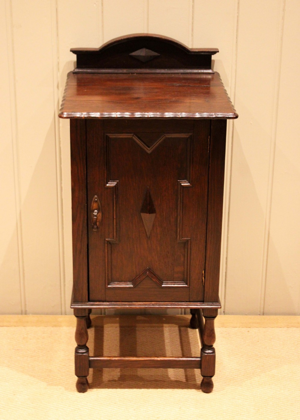 A dark oak bedside cabinet with a geometric panelled door, turned legs, low stretchers, and an arched gallery back. View III