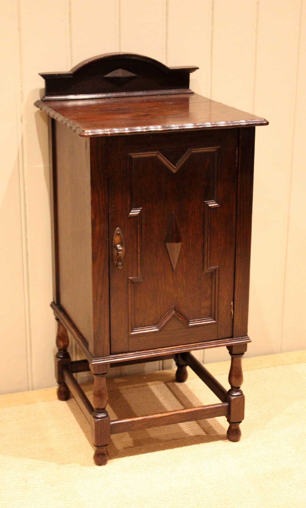 A dark oak bedside cabinet with a geometric panelled door, turned legs, low stretchers, and an arched gallery back. View IV