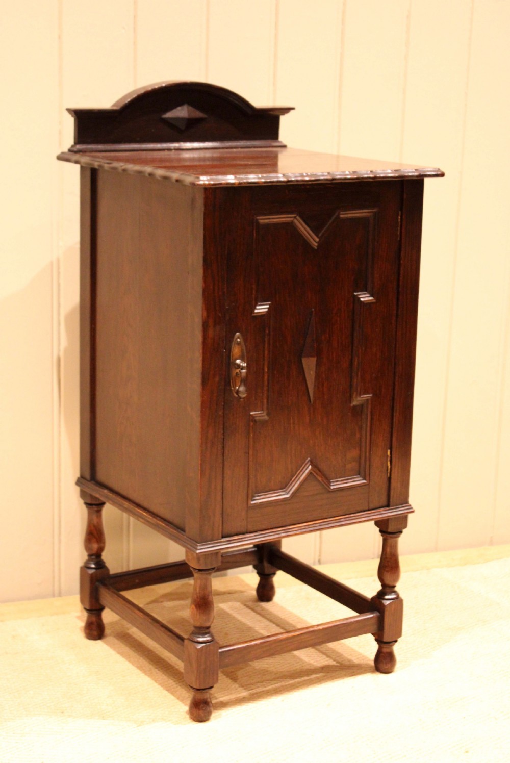 A dark oak bedside cabinet with a geometric panelled door, turned legs, low stretchers, and an arched gallery back. View VII