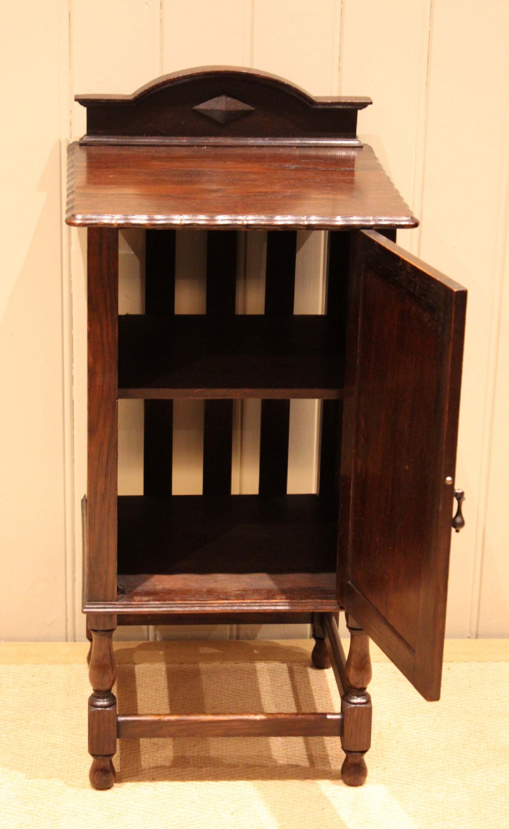 A dark oak bedside cabinet with a geometric panelled door, turned legs, low stretchers, and an arched gallery back. View VIII