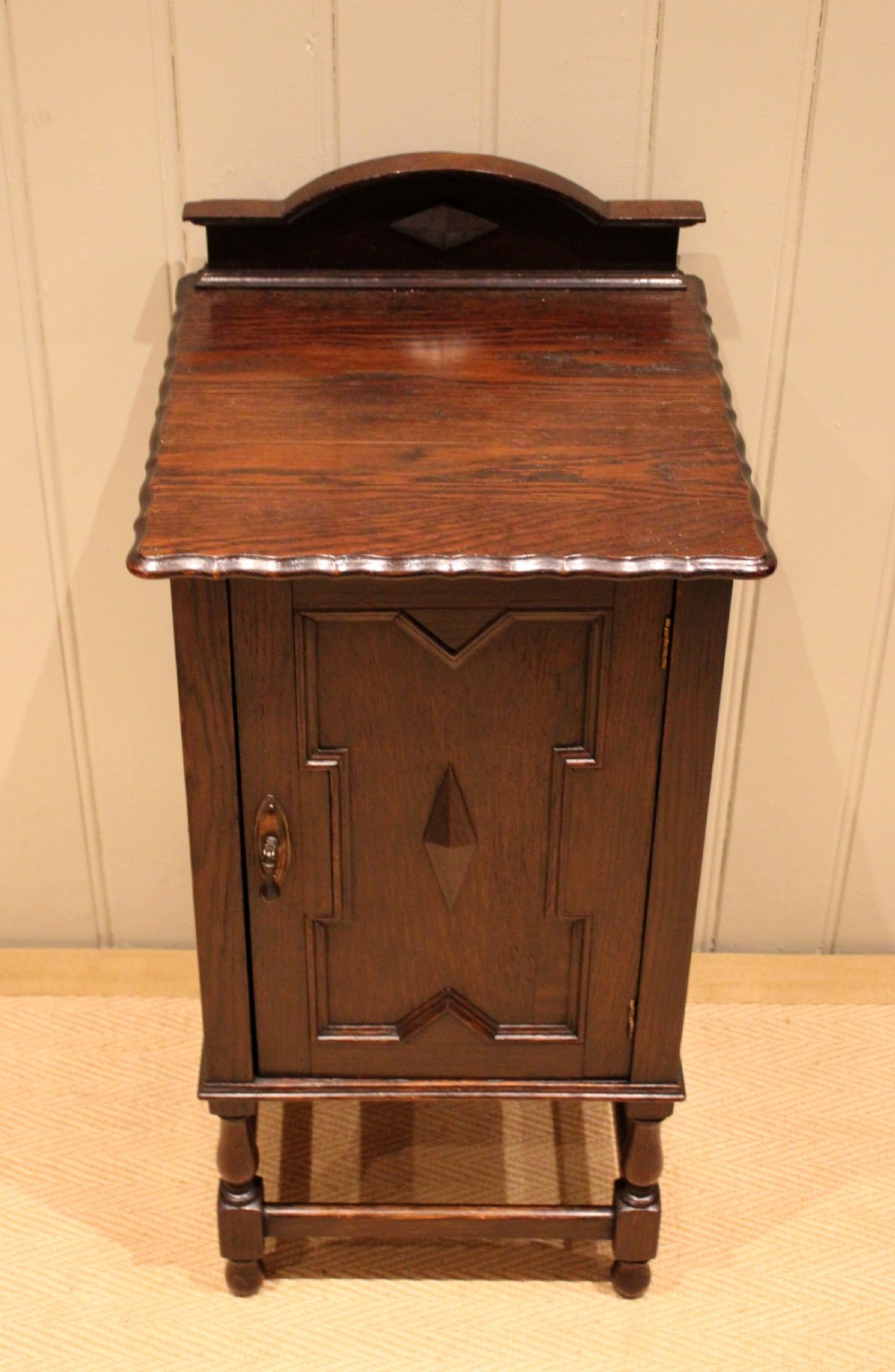A dark oak bedside cabinet with a geometric panelled door, turned legs, low stretchers, and an arched gallery back. View IX