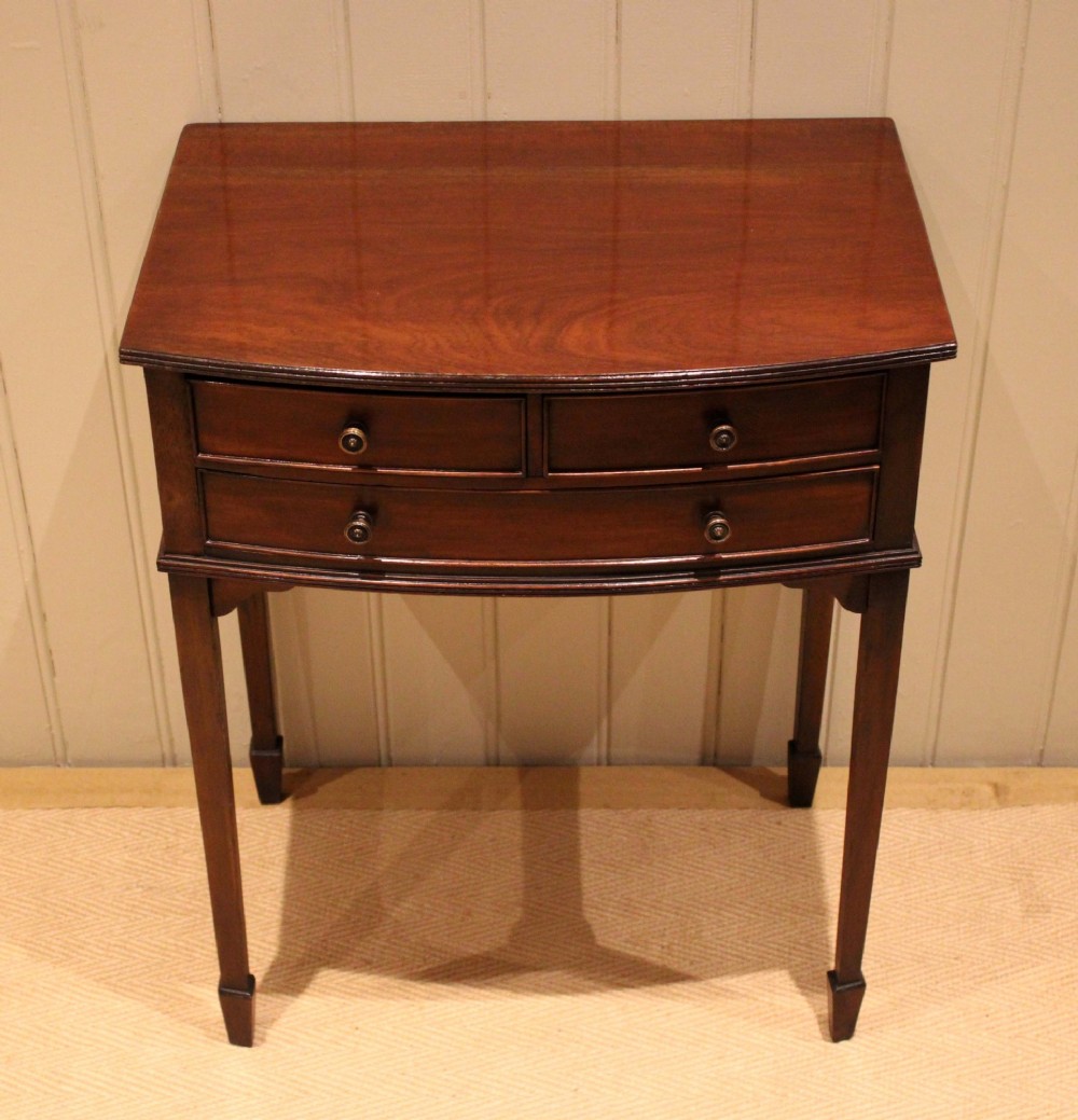 An Edwardian mahogany bow-front side table with drawers and brass handles resting on tapered legs.