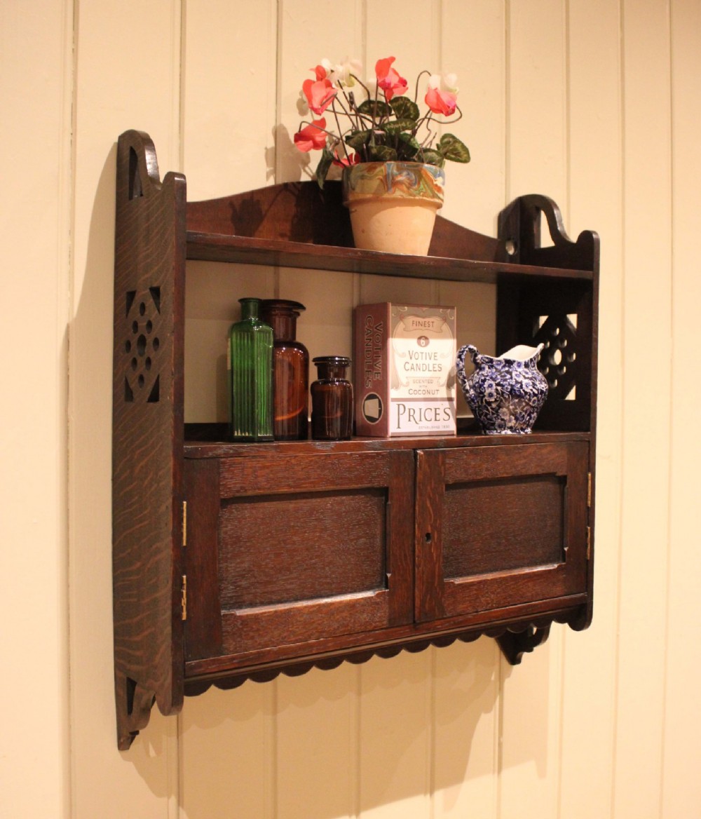 A dark oak Arts and Crafts wall-mounted cabinet with two open upper shelves above a double-door cupboard featuring pierced geometric side cut-outs and a scalloped lower trim.
