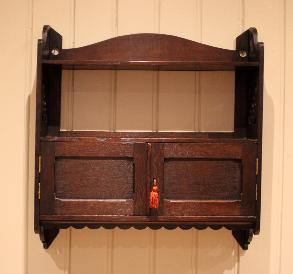 A dark oak Arts and Crafts wall-mounted cabinet with two open upper shelves above a double-door cupboard featuring pierced geometric side cut-outs and a scalloped lower trim. View II