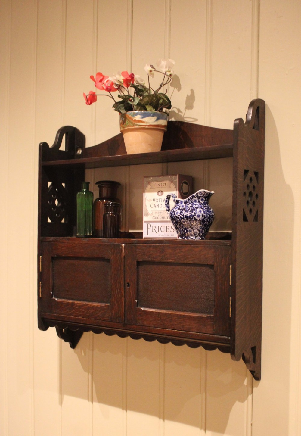 A dark oak Arts and Crafts wall-mounted cabinet with two open upper shelves above a double-door cupboard featuring pierced geometric side cut-outs and a scalloped lower trim. View III