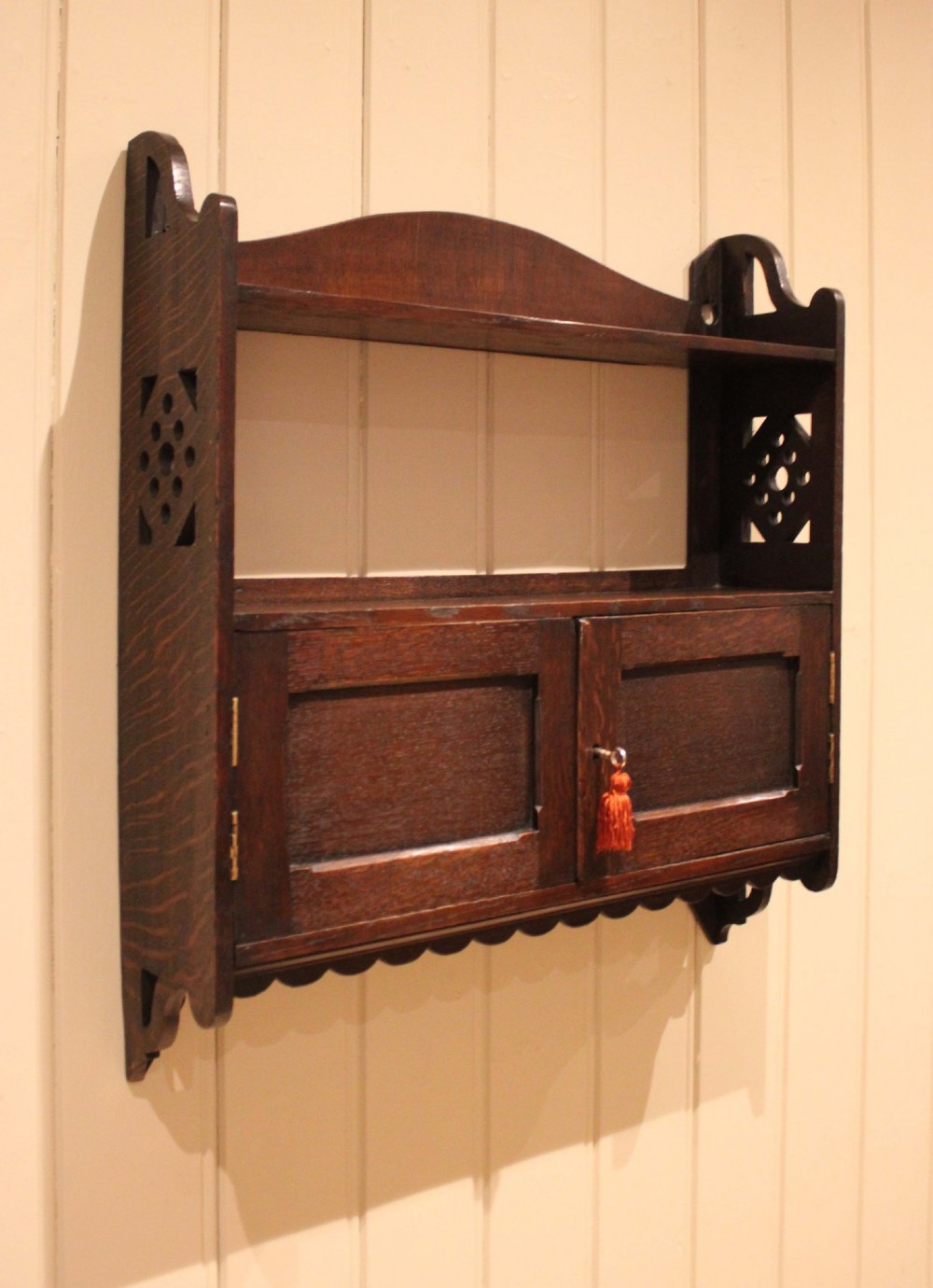 A dark oak Arts and Crafts wall-mounted cabinet with two open upper shelves above a double-door cupboard featuring pierced geometric side cut-outs and a scalloped lower trim. View IV