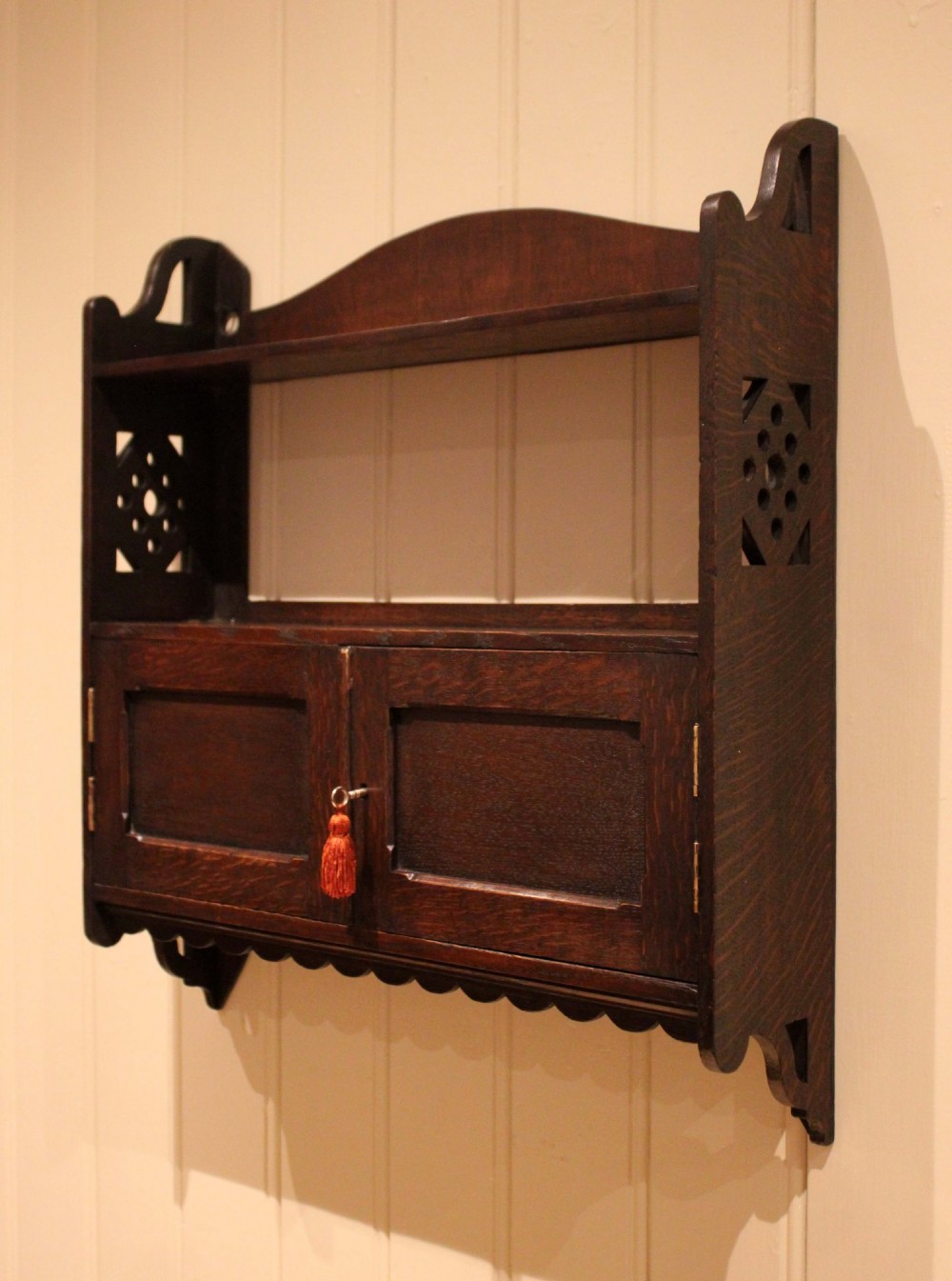 A dark oak Arts and Crafts wall-mounted cabinet with two open upper shelves above a double-door cupboard featuring pierced geometric side cut-outs and a scalloped lower trim. View VI