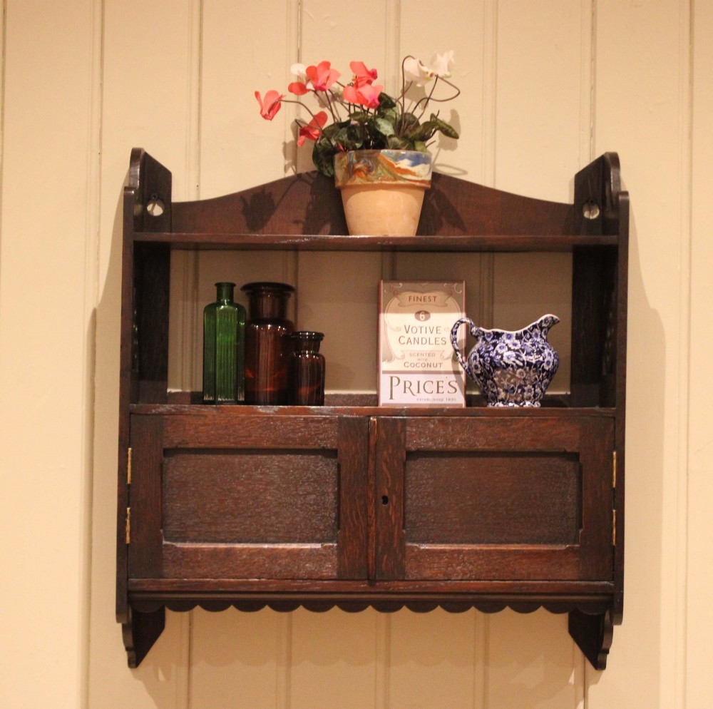 A dark oak Arts and Crafts wall-mounted cabinet with two open upper shelves above a double-door cupboard featuring pierced geometric side cut-outs and a scalloped lower trim. View VII