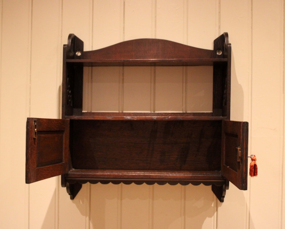 A dark oak Arts and Crafts wall-mounted cabinet with two open upper shelves above a double-door cupboard featuring pierced geometric side cut-outs and a scalloped lower trim. View VIII
