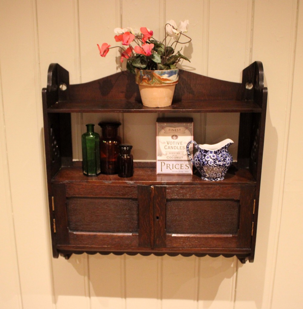 A dark oak Arts and Crafts wall-mounted cabinet with two open upper shelves above a double-door cupboard featuring pierced geometric side cut-outs and a scalloped lower trim. View IX
