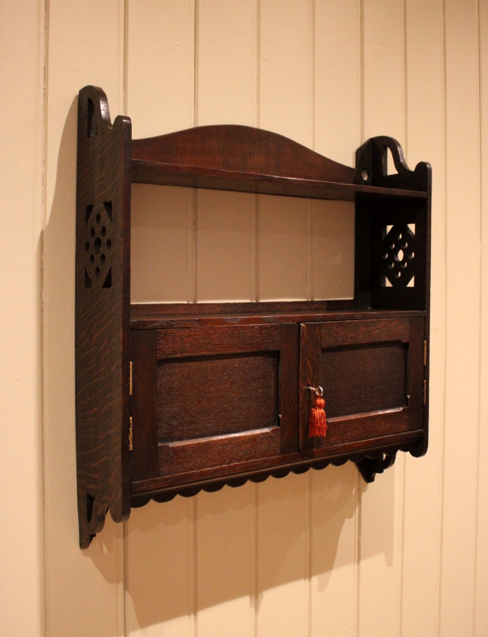 A dark oak Arts and Crafts wall-mounted cabinet with two open upper shelves above a double-door cupboard featuring pierced geometric side cut-outs and a scalloped lower trim. View X