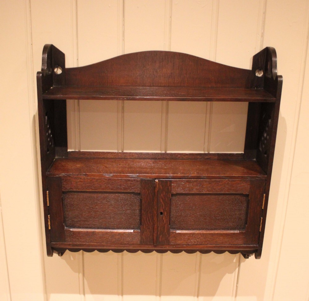 A dark oak Arts and Crafts wall-mounted cabinet with two open upper shelves above a double-door cupboard featuring pierced geometric side cut-outs and a scalloped lower trim. View XI