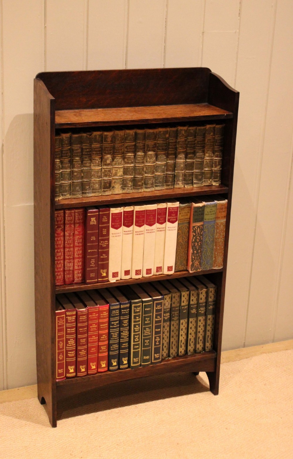A small dark oak open bookcase with three horizontal shelves filled with vintage books and a shaped top gallery.