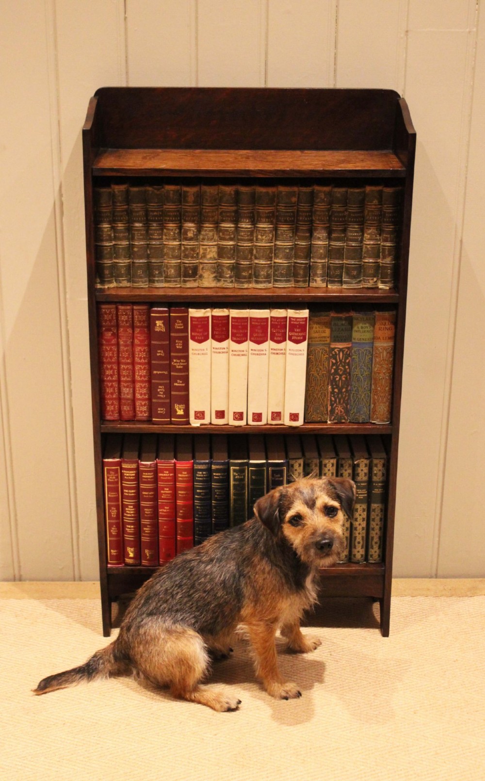 A small dark oak open bookcase with three horizontal shelves filled with vintage books and a shaped top gallery. View III