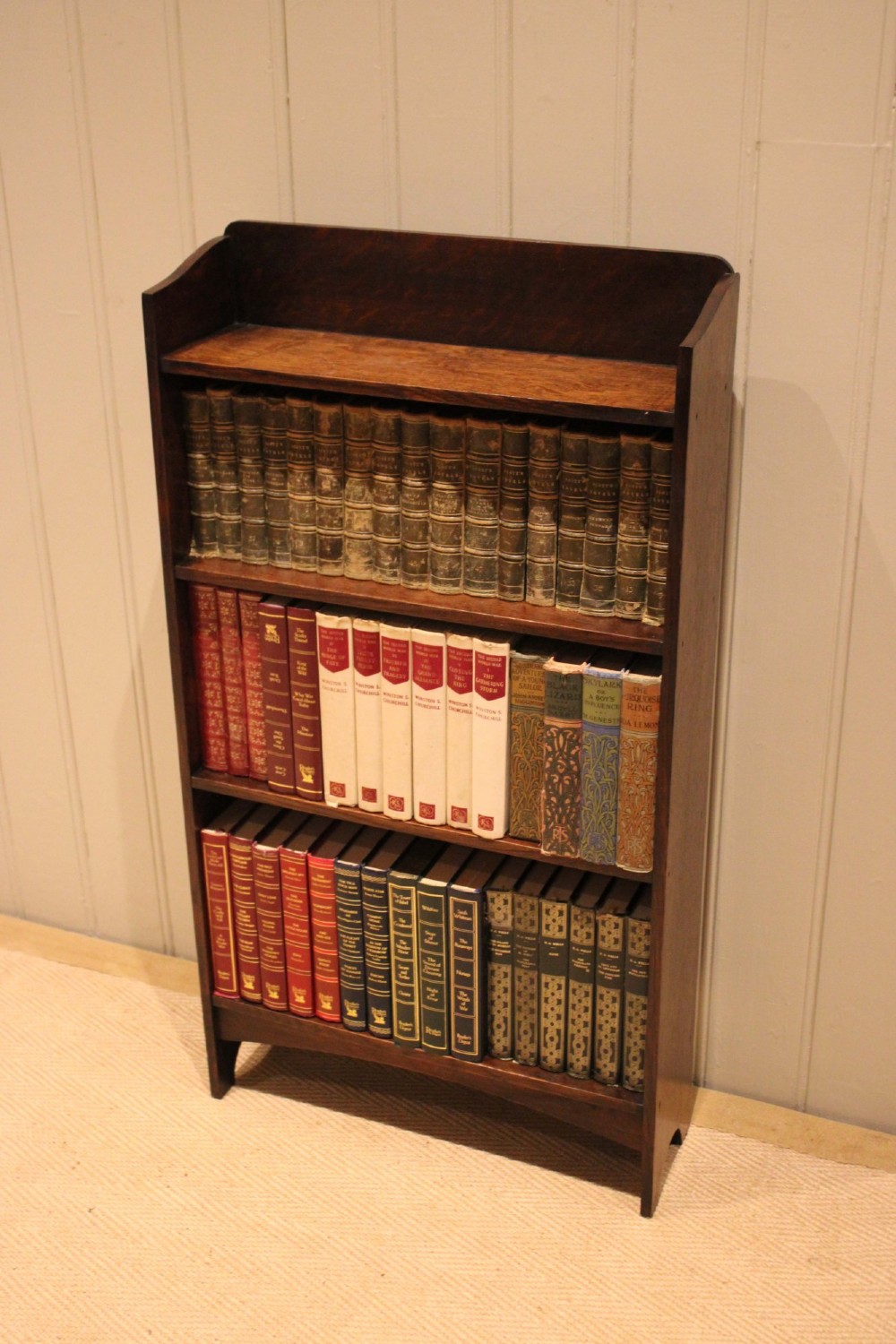 A small dark oak open bookcase with three horizontal shelves filled with vintage books and a shaped top gallery. View V