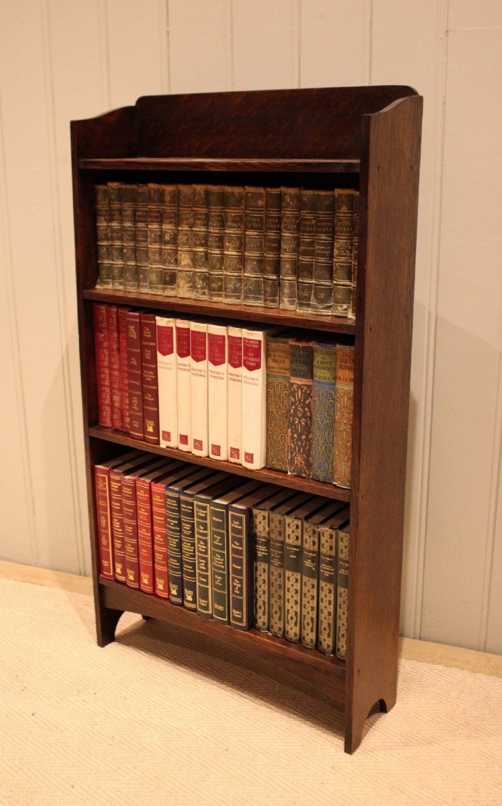 A small dark oak open bookcase with three horizontal shelves filled with vintage books and a shaped top gallery. View IX