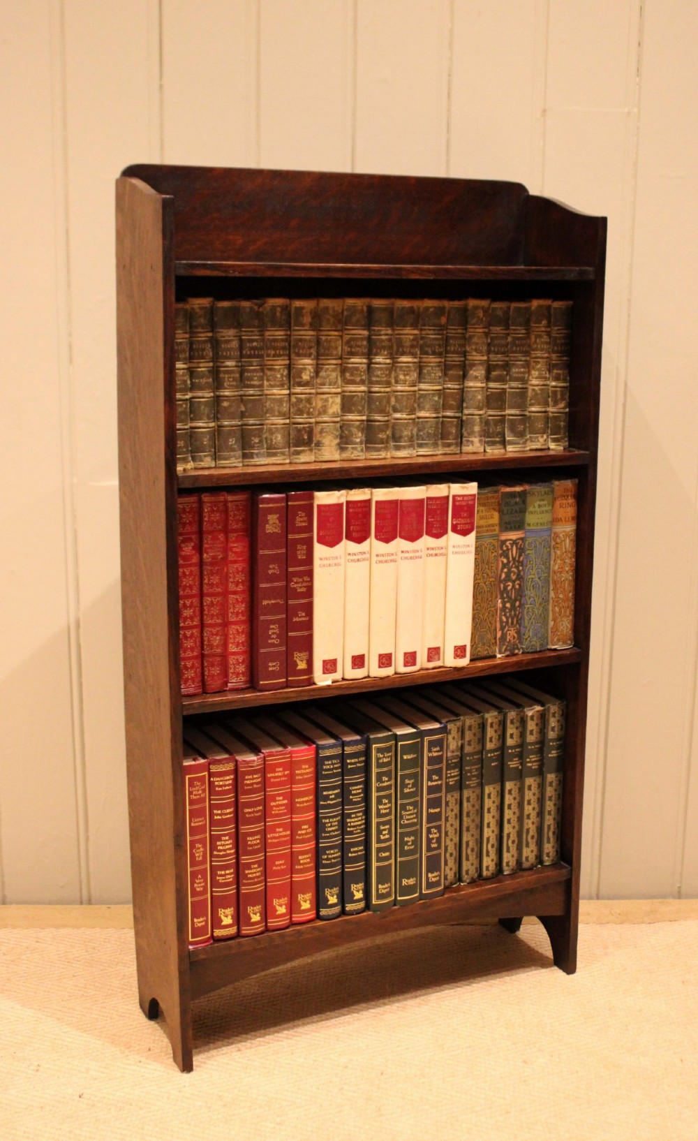 A small dark oak open bookcase with three horizontal shelves filled with vintage books and a shaped top gallery. View XI