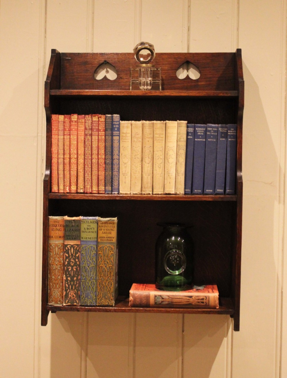Dark oak Arts and Crafts three-tier wall shelving unit with two pierced heart cut-outs along the upper gallery rail.