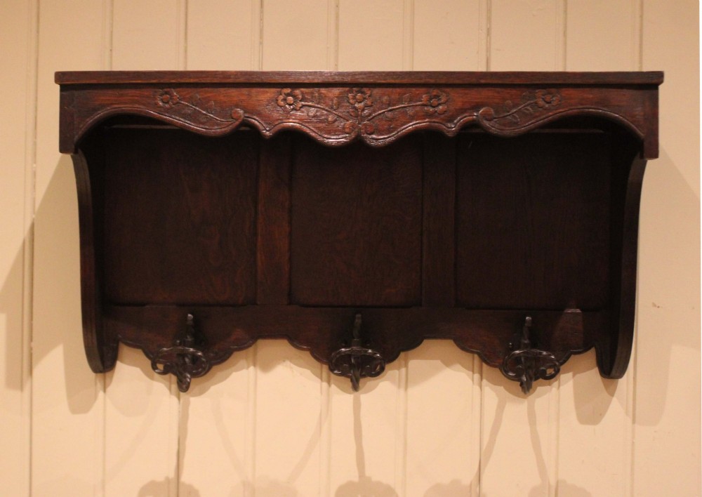 A dark oak wall-mounted coat rack featuring a top shelf, a carved floral apron, a three-panel backboard, and three metal coat hooks.