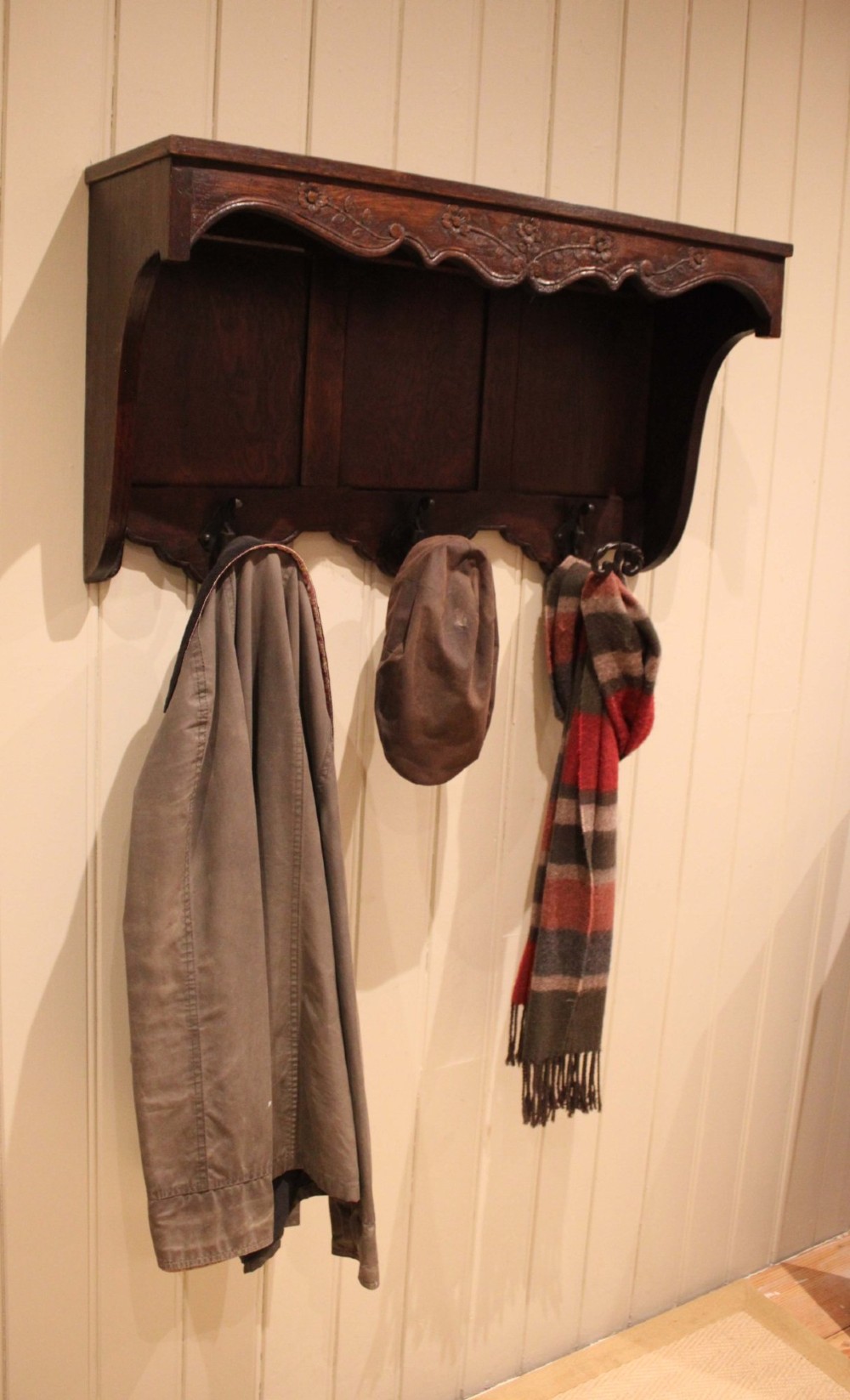 A dark oak wall-mounted coat rack featuring a top shelf, a carved floral apron, a three-panel backboard, and three metal coat hooks. View III