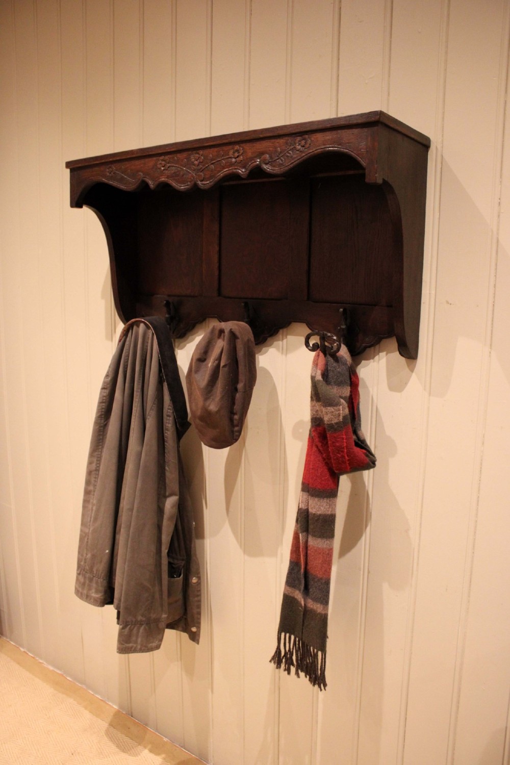 A dark oak wall-mounted coat rack featuring a top shelf, a carved floral apron, a three-panel backboard, and three metal coat hooks. View IV