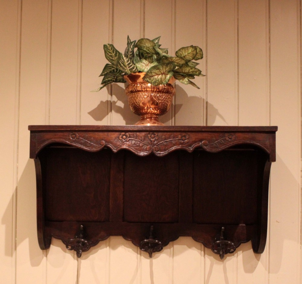 A dark oak wall-mounted coat rack featuring a top shelf, a carved floral apron, a three-panel backboard, and three metal coat hooks. View V