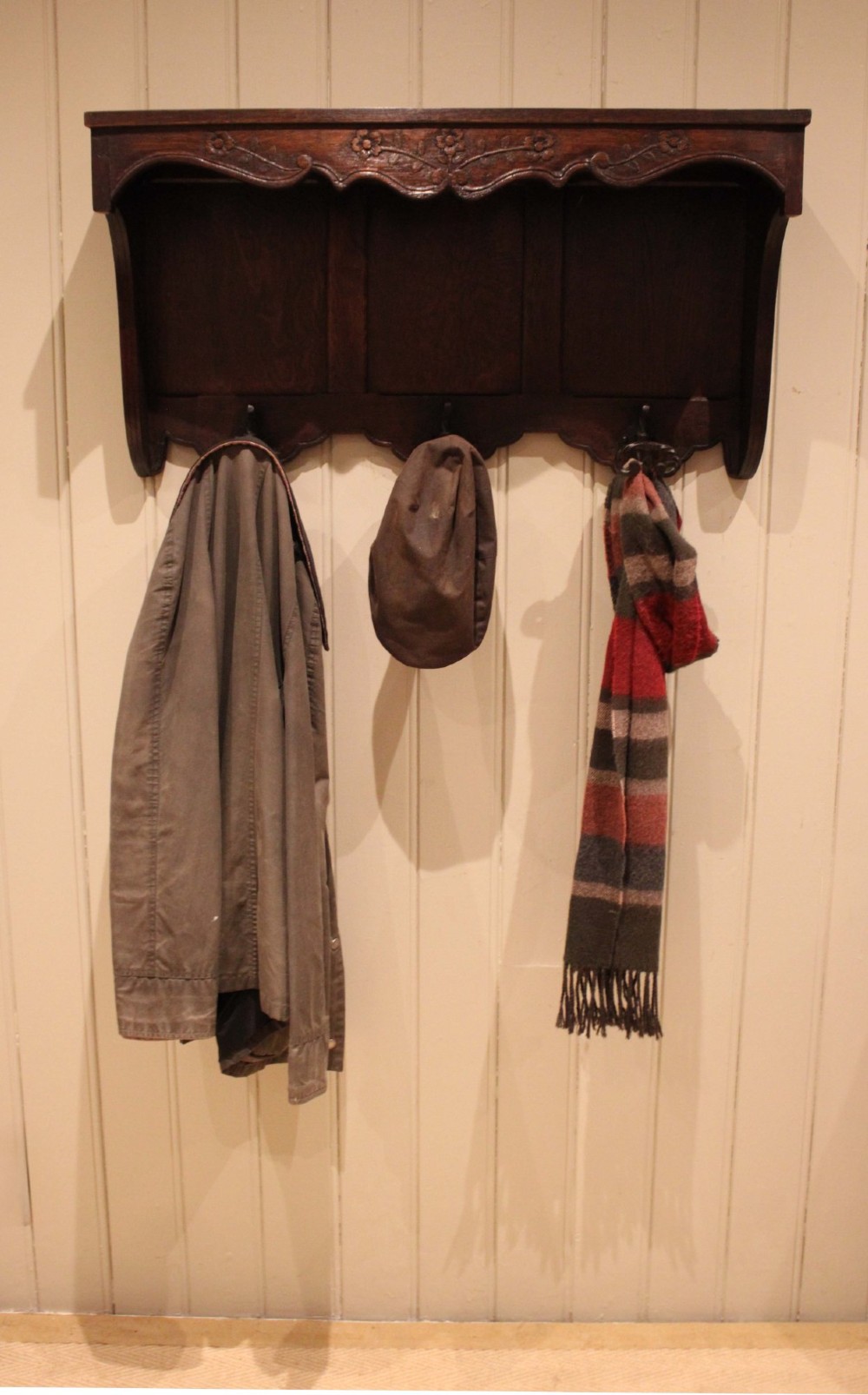 A dark oak wall-mounted coat rack featuring a top shelf, a carved floral apron, a three-panel backboard, and three metal coat hooks. View VI