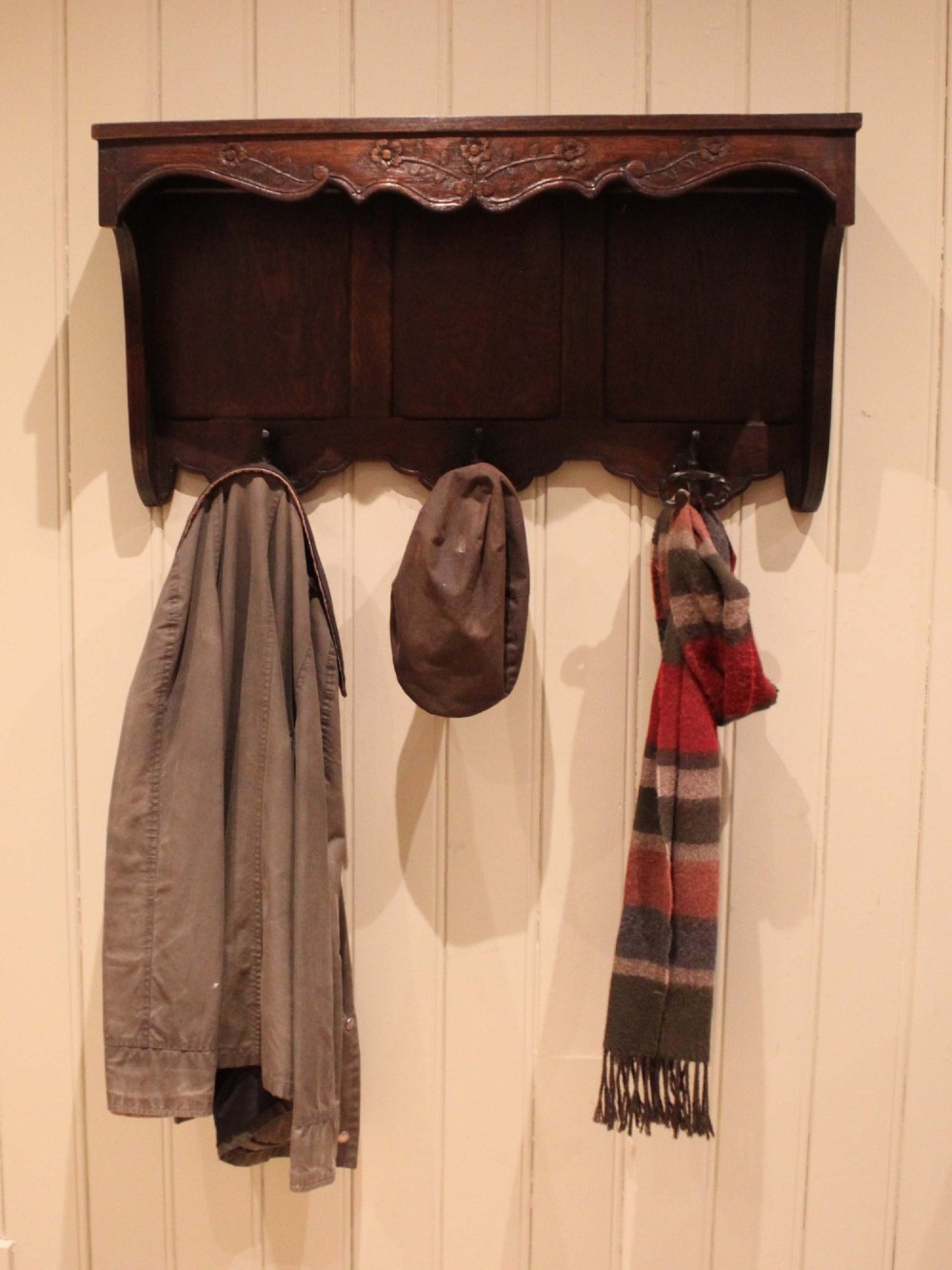 A dark oak wall-mounted coat rack featuring a top shelf, a carved floral apron, a three-panel backboard, and three metal coat hooks. View VII