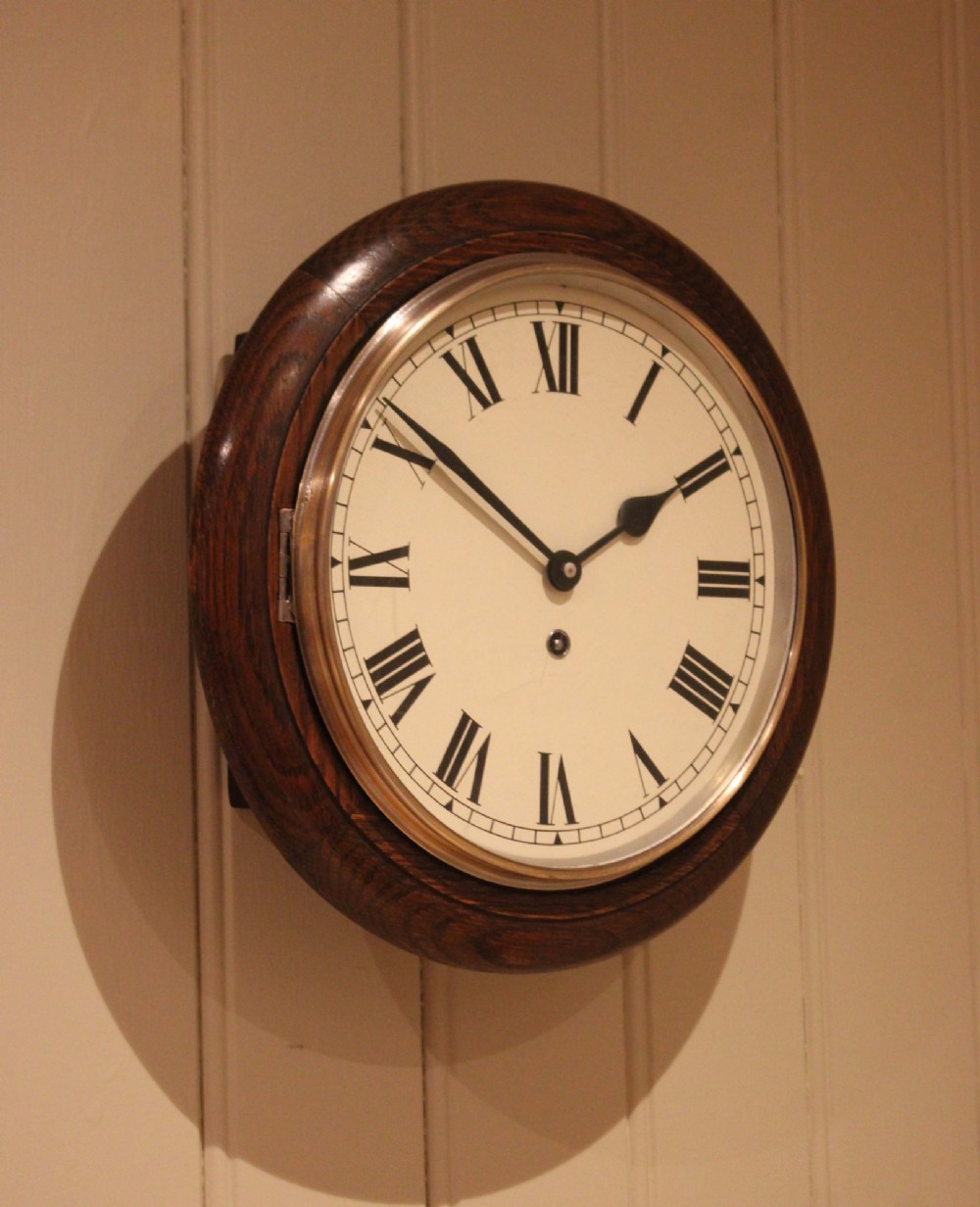 A circular early 20th-century wall clock featuring a moulded dark oak frame, a brass bezel, and a white dial face with black Roman numerals.
