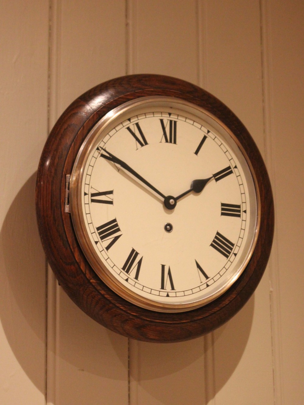 A circular early 20th-century wall clock featuring a moulded dark oak frame, a brass bezel, and a white dial face with black Roman numerals. View IV