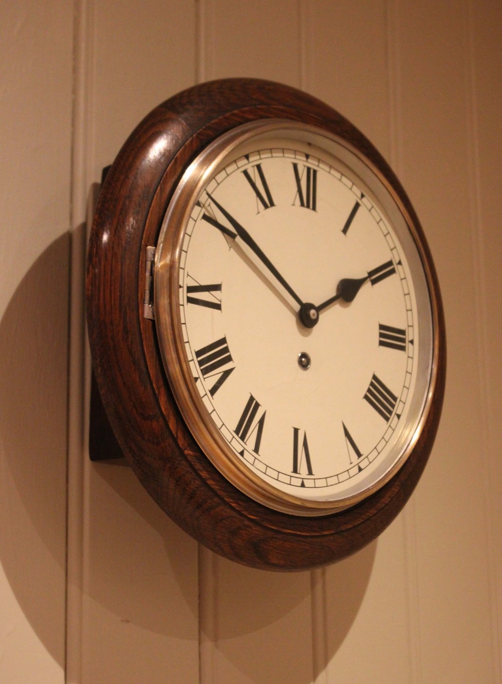 A circular early 20th-century wall clock featuring a moulded dark oak frame, a brass bezel, and a white dial face with black Roman numerals. View V